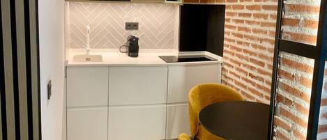 A modern kitchen with white cabinetry, a built-in oven, and a black range hood. There is a yellow chair and a small round table in the dining area.