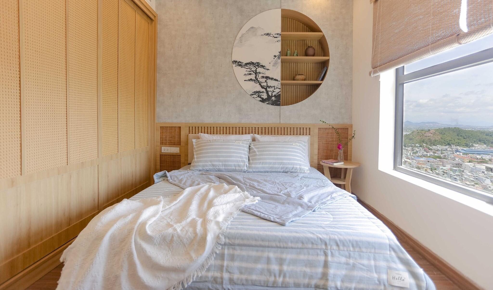 A bedroom with a bed, a round mirror with a painting, a small table with a vase, and a window with a view of a cityscape.