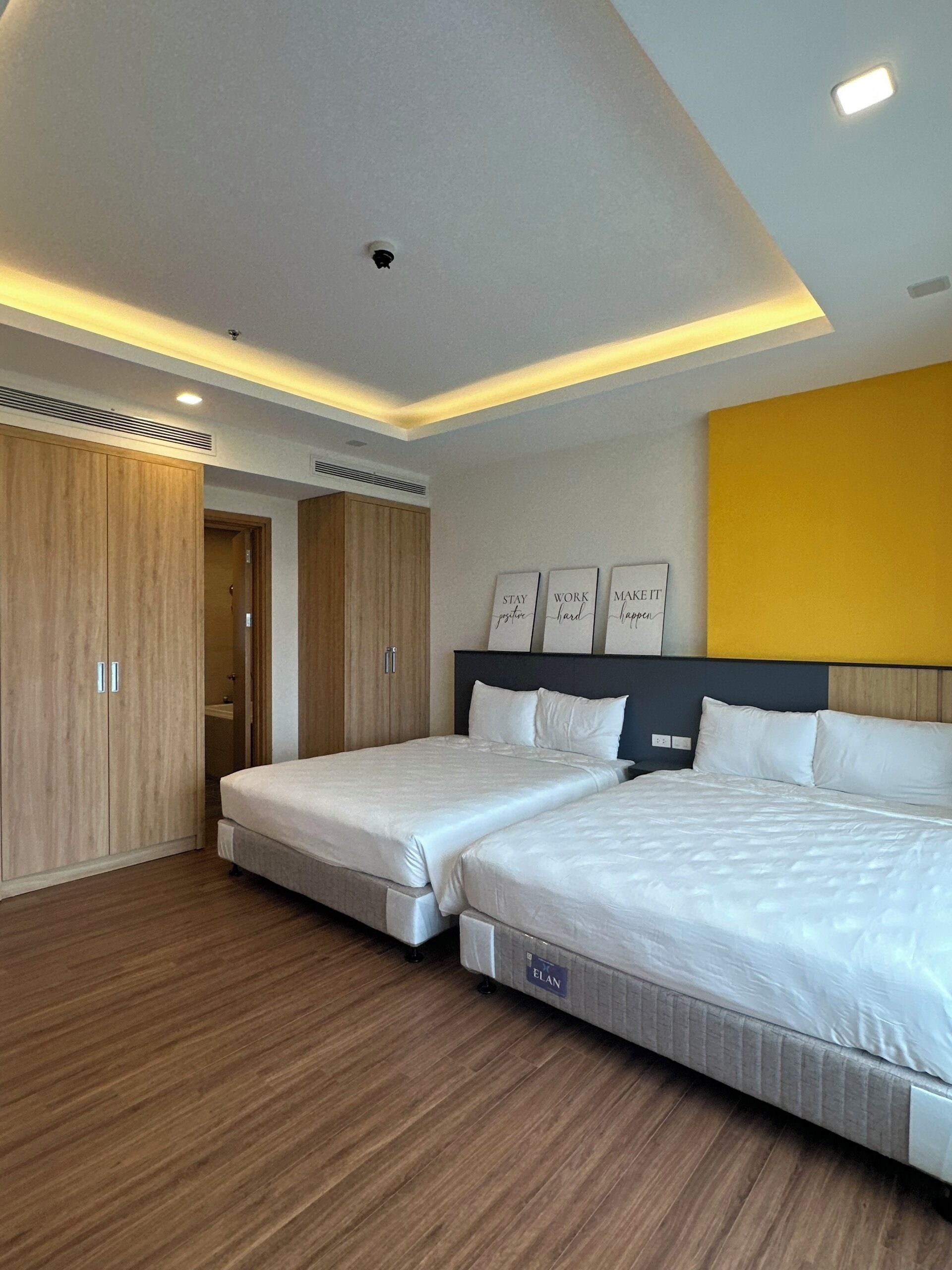 Quarto de hotel moderno com duas camas, um guarda-roupa de madeira e arte motivacional na parede.
