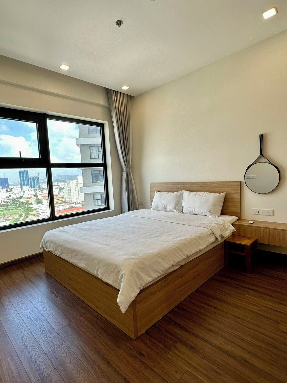 Una habitación de hotel moderna con una cama grande, piso de madera, vistas a la ciudad y una mesita de noche sencilla.