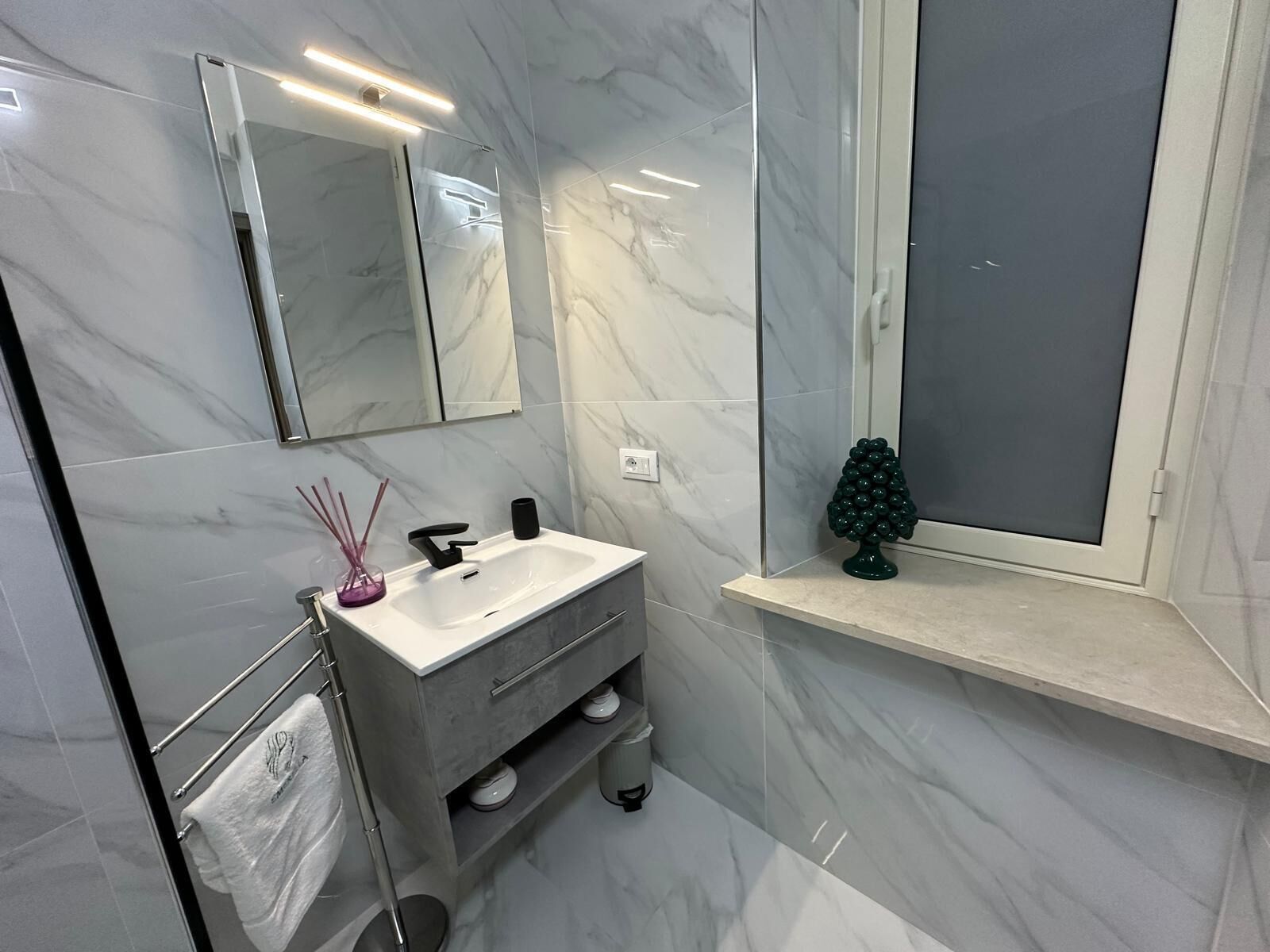 Une salle de bain avec un sol en marbre, un lavabo blanc avec un robinet noir, une petite étagère ornée d’un objet décoratif et un porte-serviettes avec une serviette blanche.