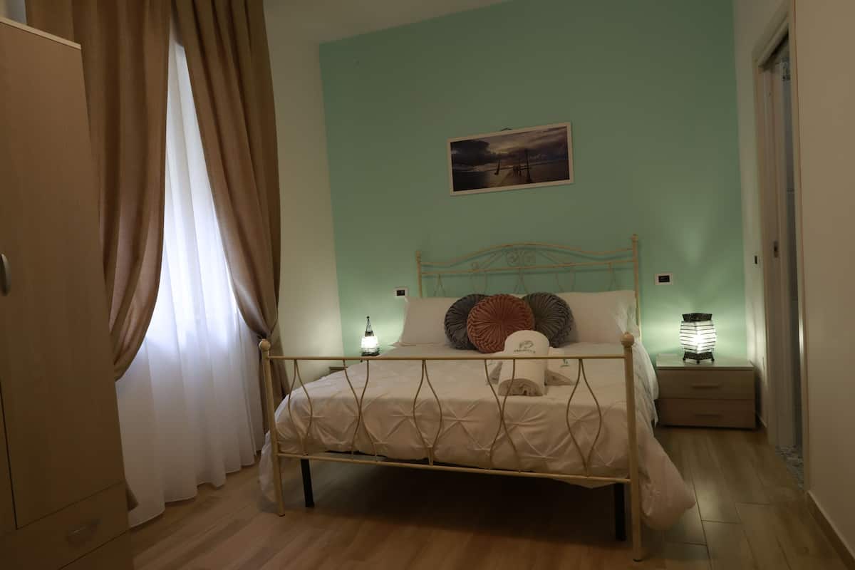 A bedroom with a bed, curtains, a nightstand, a wall-mounted lamp, and a painting.