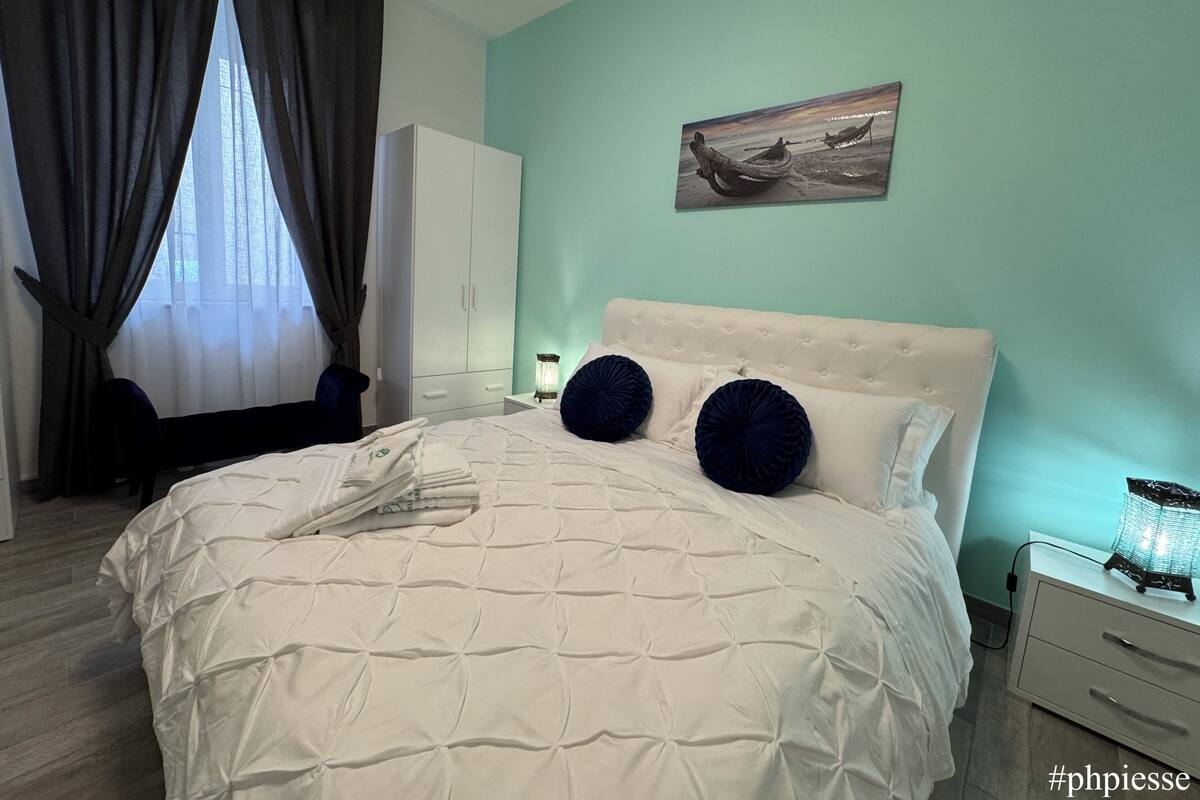 A neatly made bed with a white duvet and two dark blue pillows, a white wardrobe, a small bedside table with a lamp, and a painting on the wall.