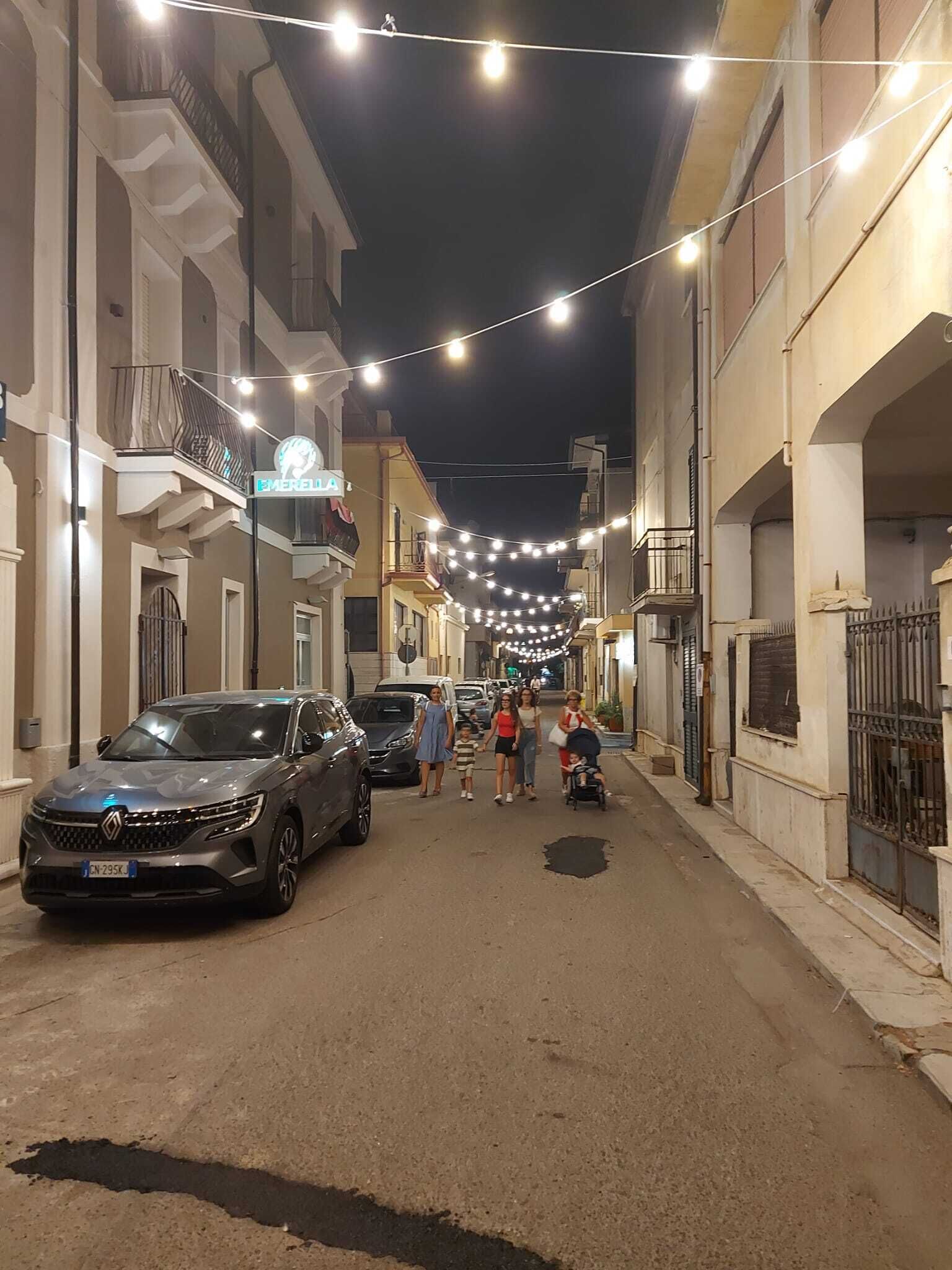 Una strada fiancheggiata da edifici, con luci decorative appese e auto parcheggiate.