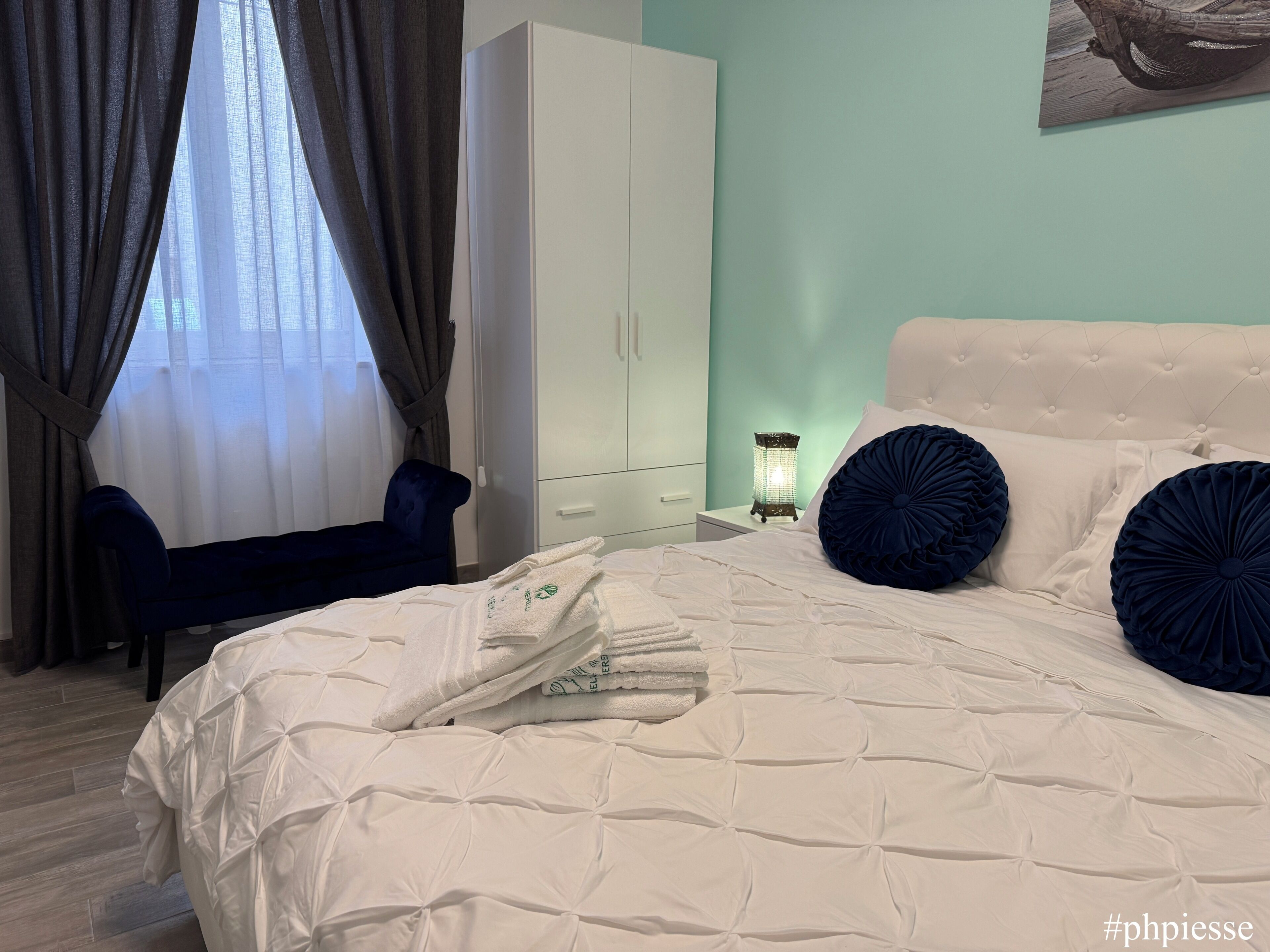 Una cama bien hecha con ropa de cama blanca y dos almohadas azul oscuro, un armario blanco, una silla azul y una mesita con una vela encendida.