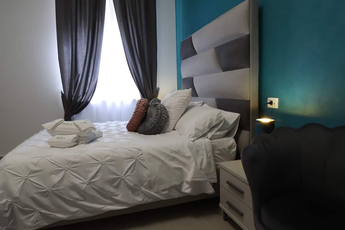 A neatly made bed with a white duvet, pillows, and a folded towel. There is a bedside table with a lamp, a dark-colored chair, and a wall with a power outlet.