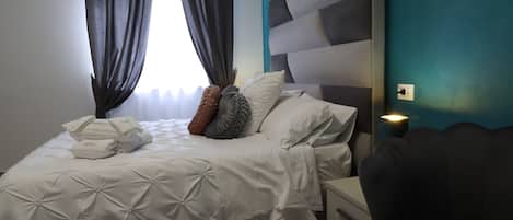 A neatly made bed with a white duvet, pillows, and a folded towel. There is a bedside table with a lamp, a dark-colored chair, and a wall with a power outlet.