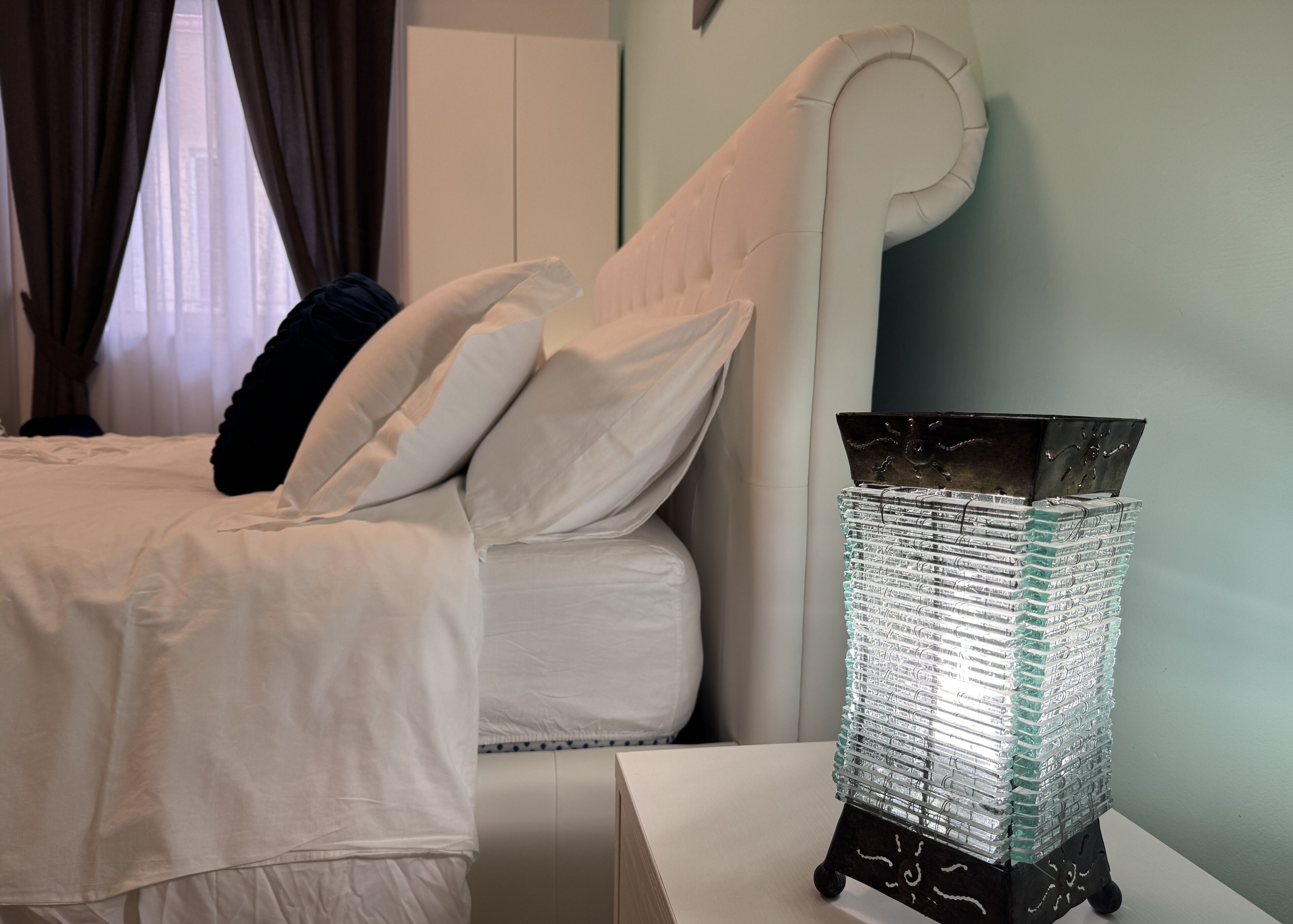 Una cama bien hecha con sábanas y almohadas blancas, una moderna mesita de noche con un jarrón de vidrio decorativo, y vistas a un paisaje urbano a través de cortinas translúcidas.
