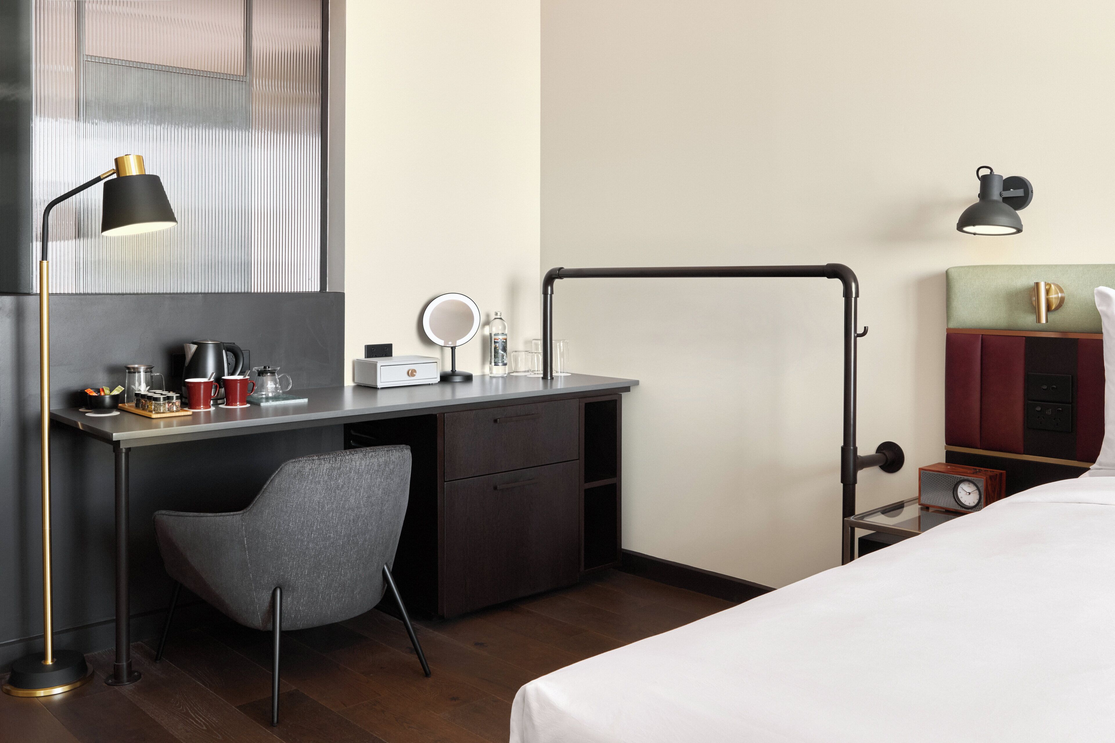 A hotel room with a bed, a desk with a chair, a lamp, a mirror, a clock, and a wardrobe.