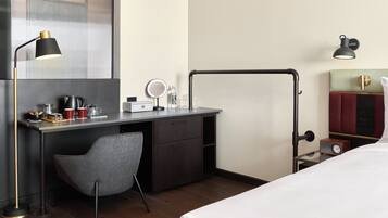 A hotel room with a bed, a desk with a chair, a lamp, a mirror, a clock, and a wardrobe.