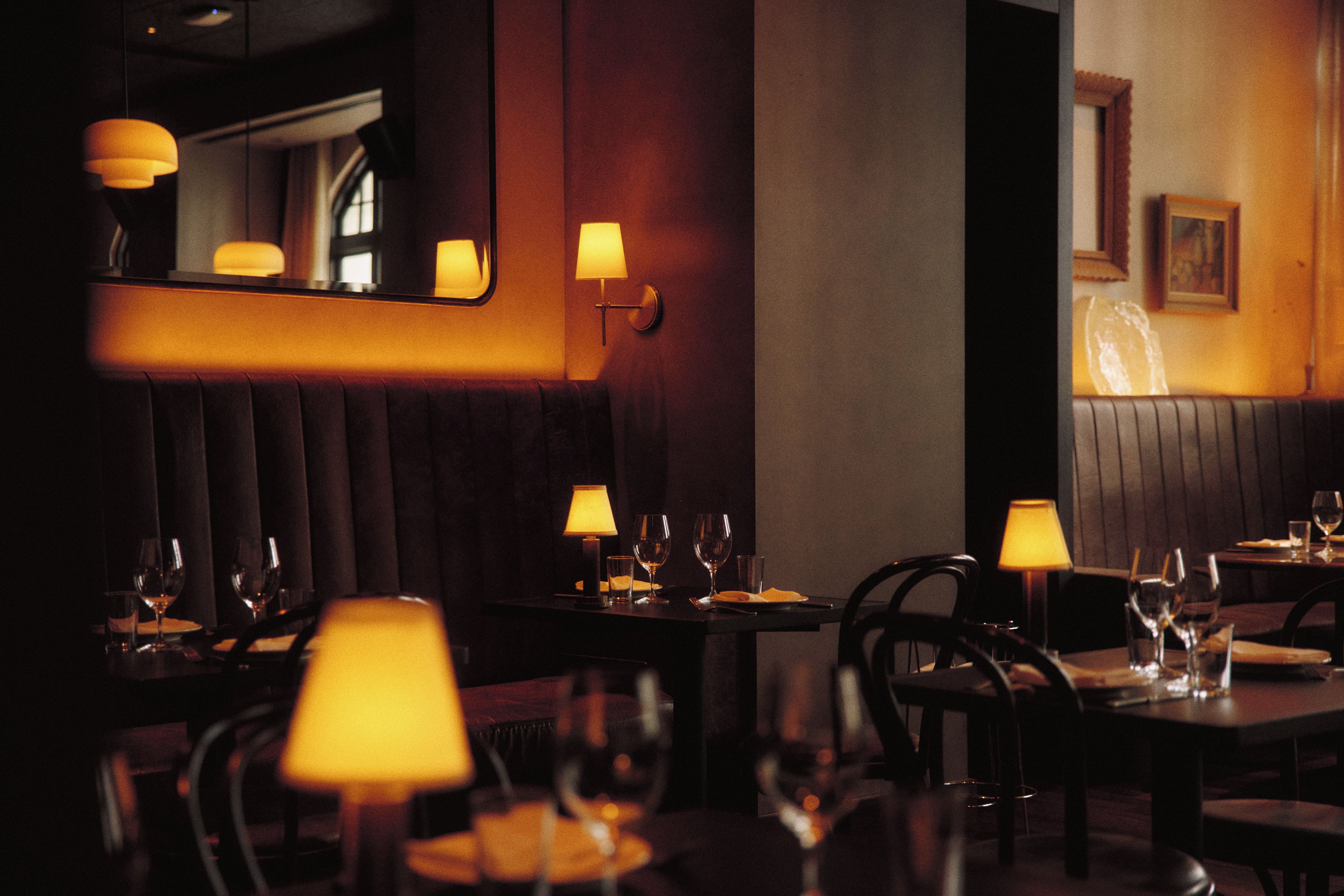 A dimly lit restaurant interior with tables set for dining, featuring cushioned booths and wall-mounted lamps.