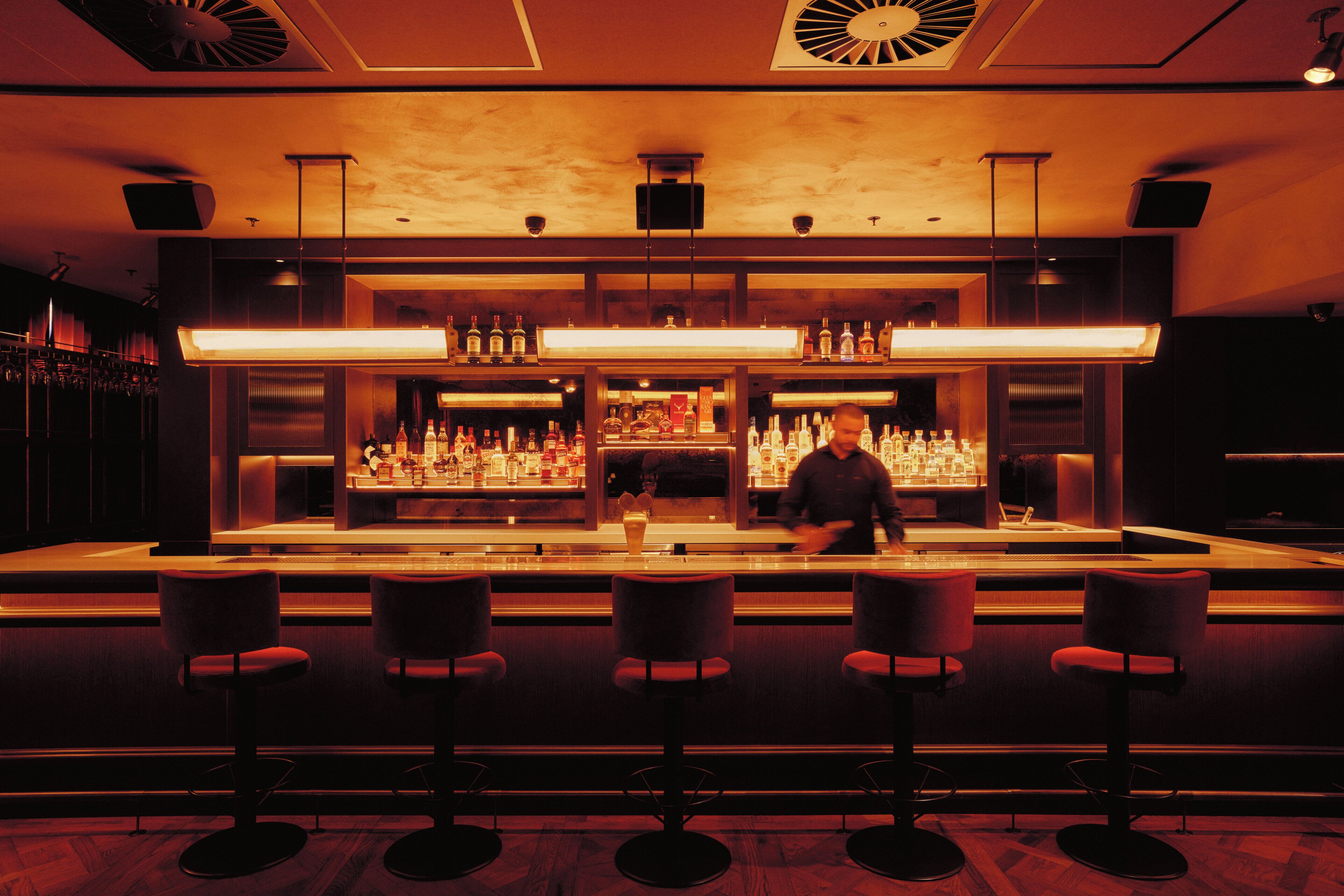 A bar area with a long counter, multiple shelves of bottles, and four stools.