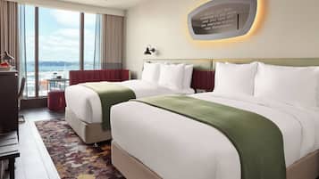 A hotel room with two beds, a large window with a city view, a quote on the wall, and a desk with a lamp.