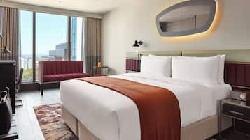 A hotel room with a large bed, a red armchair, a desk with a lamp, and a view of the city through large windows.