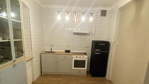 Une cuisine compacte avec des murs recouverts de carreaux blancs, un plancher en bois, un four encastré, un réfrigérateur noir et une armoire en verre dotée d’une poignée en métal.