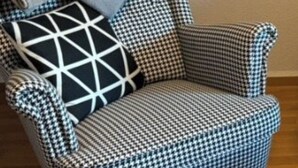 Un fauteuil à motifs à carreaux, agrémenté d’un coussin aux motifs géométriques, est placé sur un plancher en bois, adossé à un mur uni.
