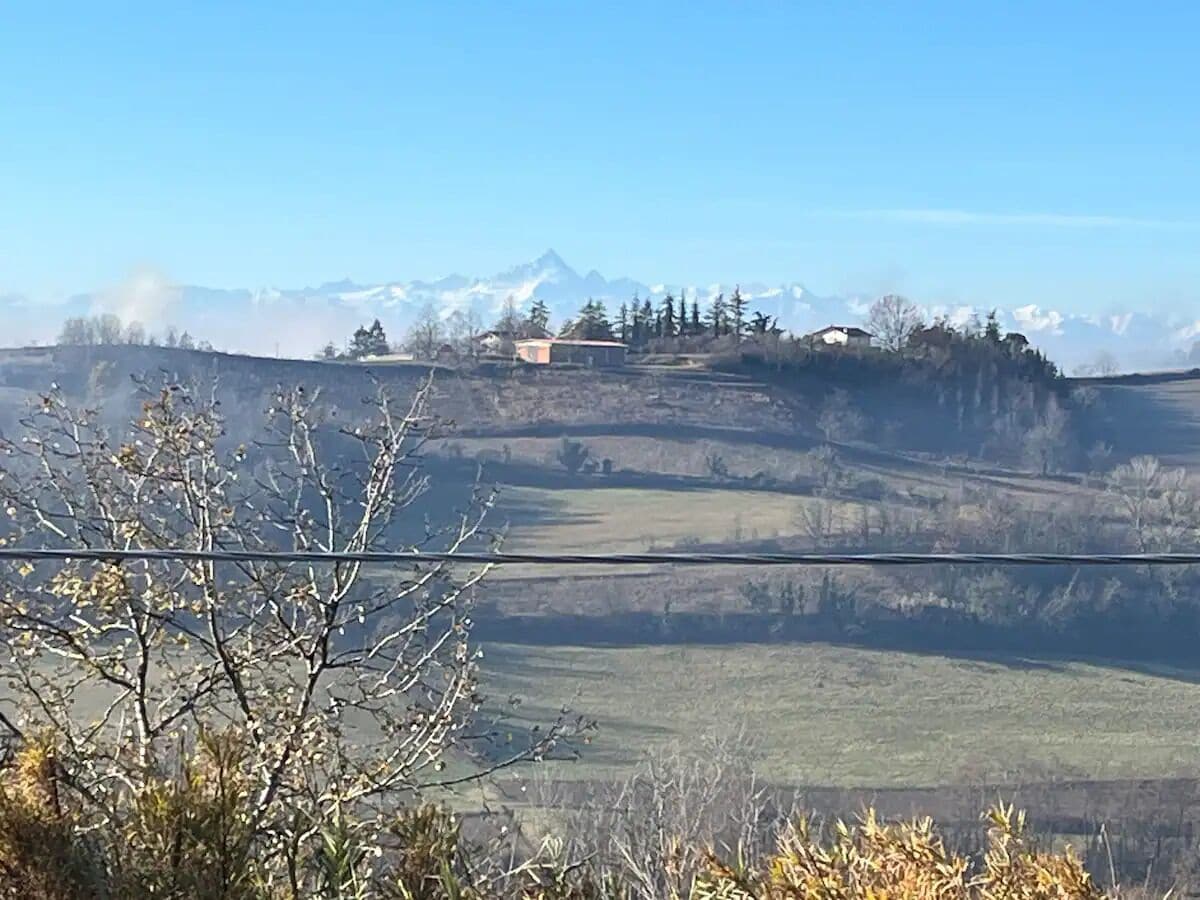 Ländliche Landschaft mit einem Haus auf einem Hügel, umgeben von Feldern und Bergen in der Ferne.