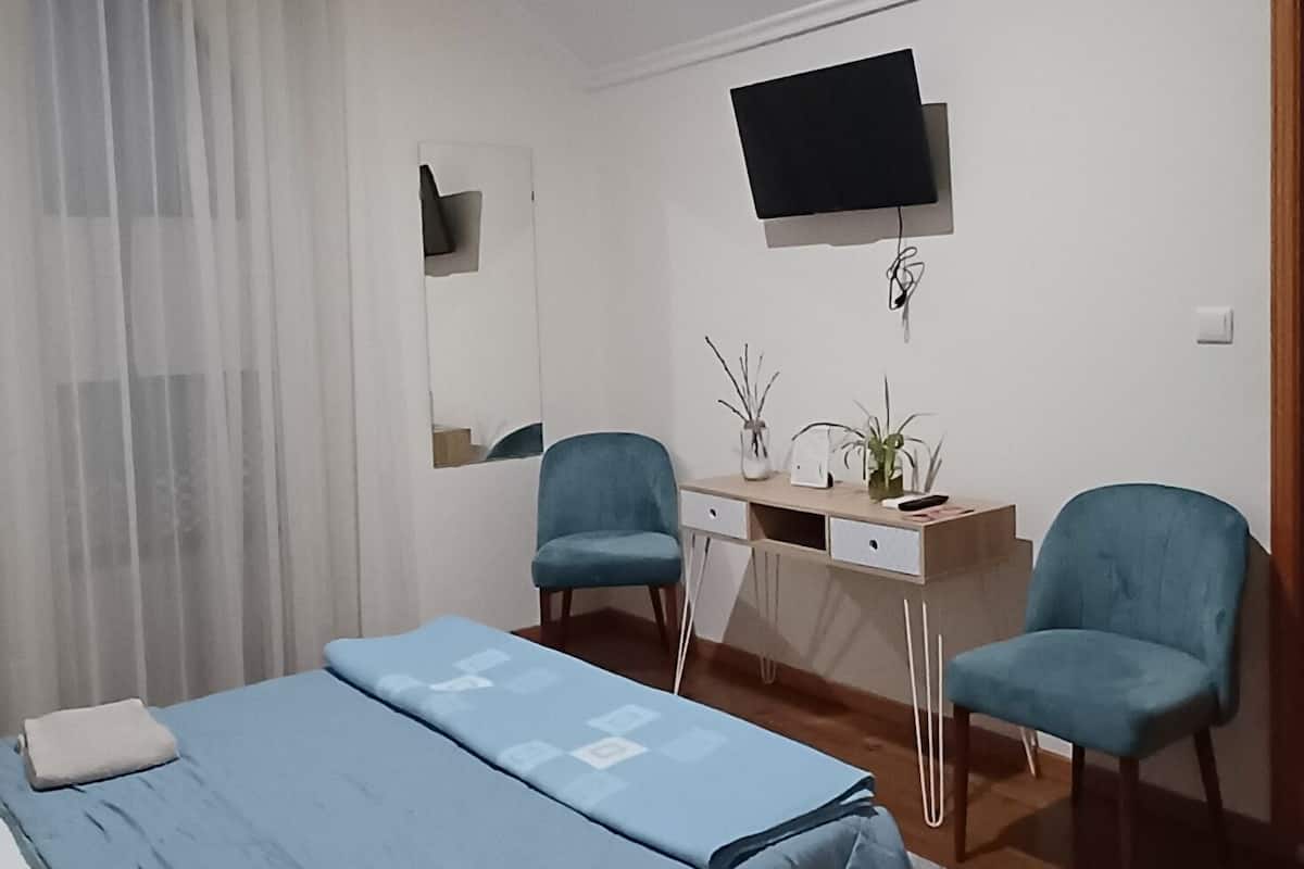 Une chambre à coucher avec un lit, deux chaises, un bureau, une télévision et une fenêtre avec des rideaux.