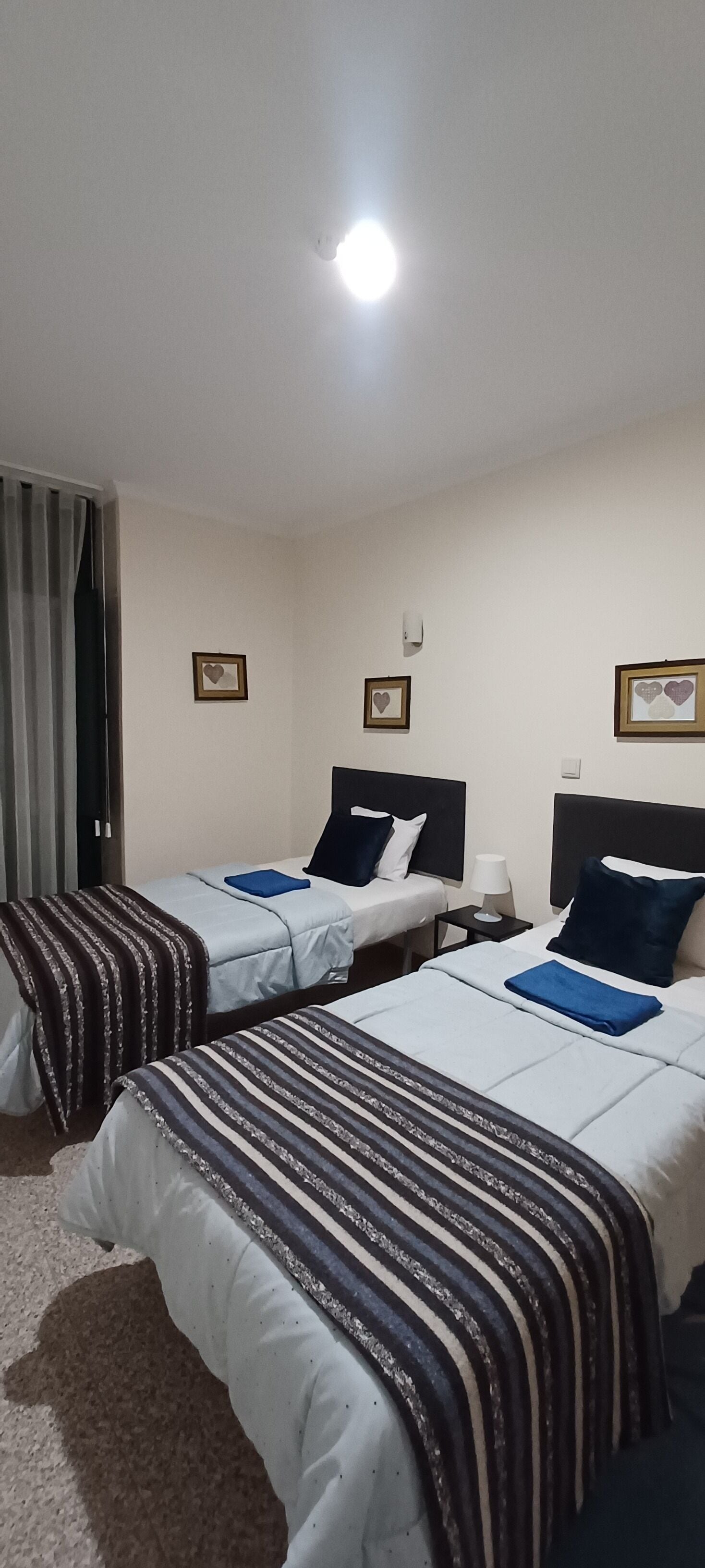 A hotel room with two single beds, each with a striped blanket and a blue pillow. There are framed pictures on the wall, a bedside table with a lamp, and a window with curtains.