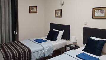 A hotel room with two single beds, each with a striped blanket and a blue pillow. There are framed pictures on the wall, a bedside table with a lamp, and a window with curtains.