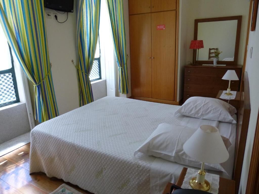 A neatly made bed with white linens, a bedside lamp, a wooden wardrobe, and a window with curtains.