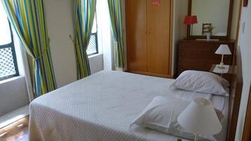 A neatly made bed with white linens, a bedside lamp, a wooden wardrobe, and a window with curtains.