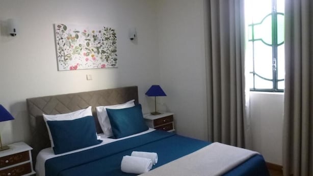 A neatly made bed with blue and white bedding, two bedside lamps, a floral artwork on the wall, and a window with curtains.