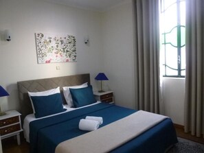 Una cama bien tendida con ropa de cama azul y blanca, dos lámparas de noche, una obra de arte floral en la pared y una ventana con cortinas.