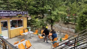 Un área de estar al aire libre con sillas y mesas anaranjadas, una estructura de madera con un letrero para un restaurante y cafetería, y un paisaje montañoso cubierto de bosque al fondo.
