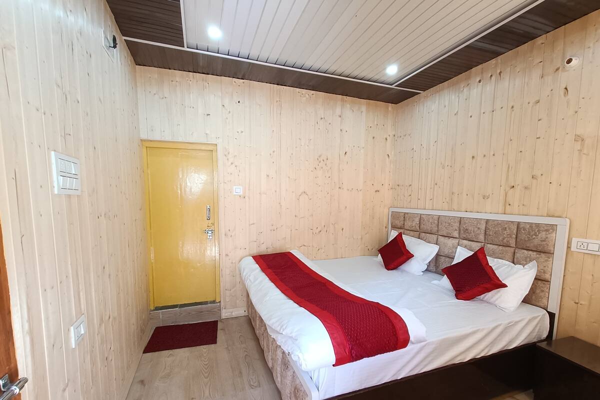 Una habitación con paredes de madera, una cama con ropa de cama blanca y detalles rojos, una puerta amarilla y una mesita de noche pequeña.