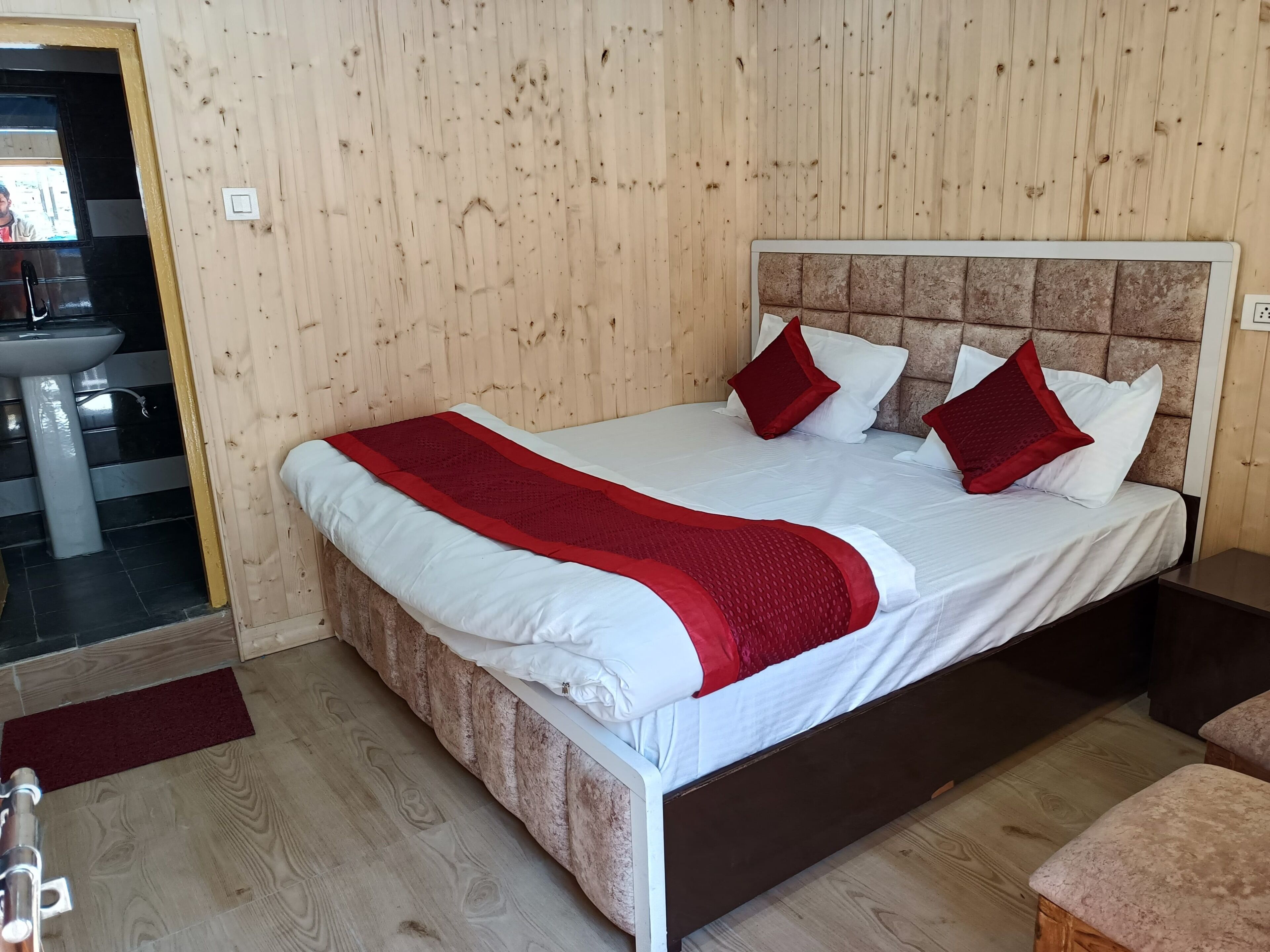 A bedroom with a wooden headboard, a bed with white and red bedding, a bedside table, and a bathroom visible through an open door.