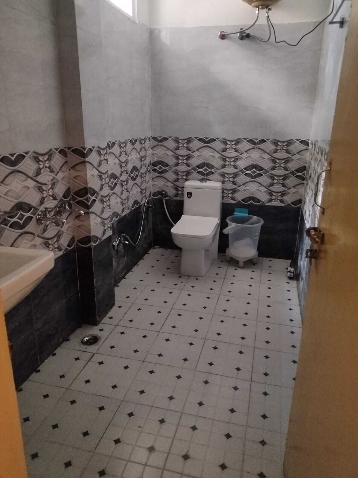 A small bathroom with a toilet, a bidet, and a sink. The walls are tiled with a decorative pattern, and the floor has a simple geometric design.