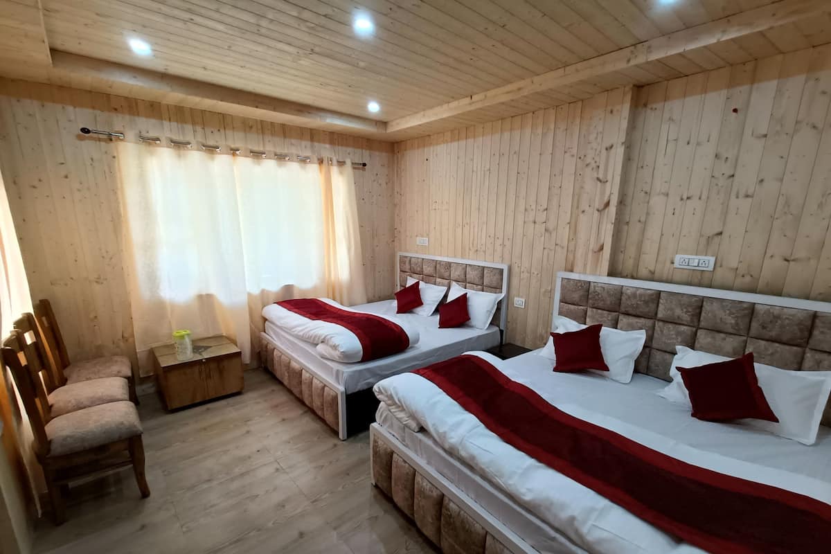 Una habitación con dos camas, pisos de madera, una silla, una mesita y un ventanal con cortinas.
