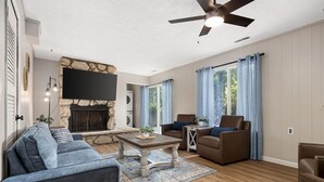 A living room with a stone fireplace, a flat-screen TV, a ceiling fan, and a large window with curtains.