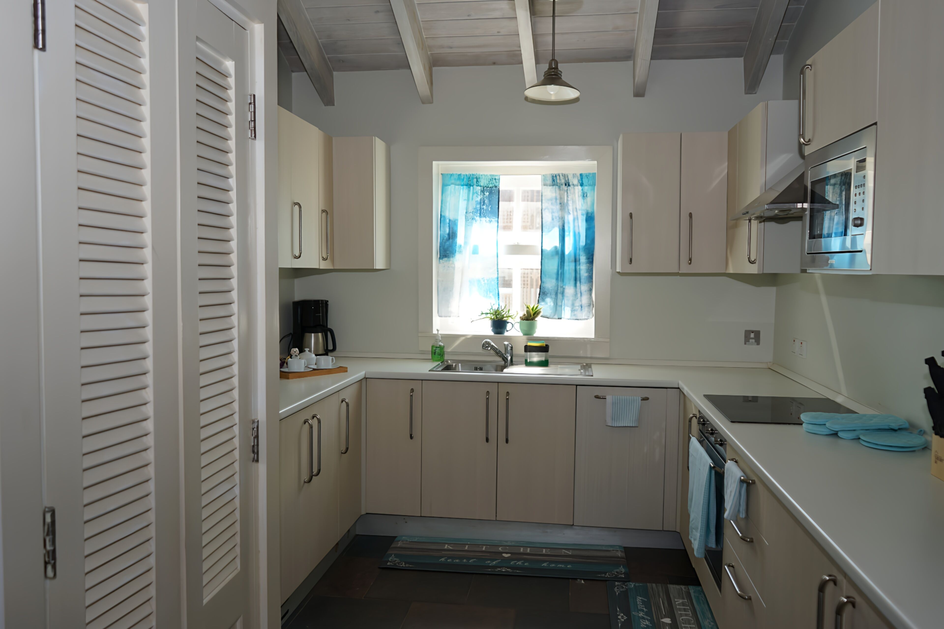 A compact kitchen with white cabinets, a built-in oven, a microwave, and a window with blue curtains.