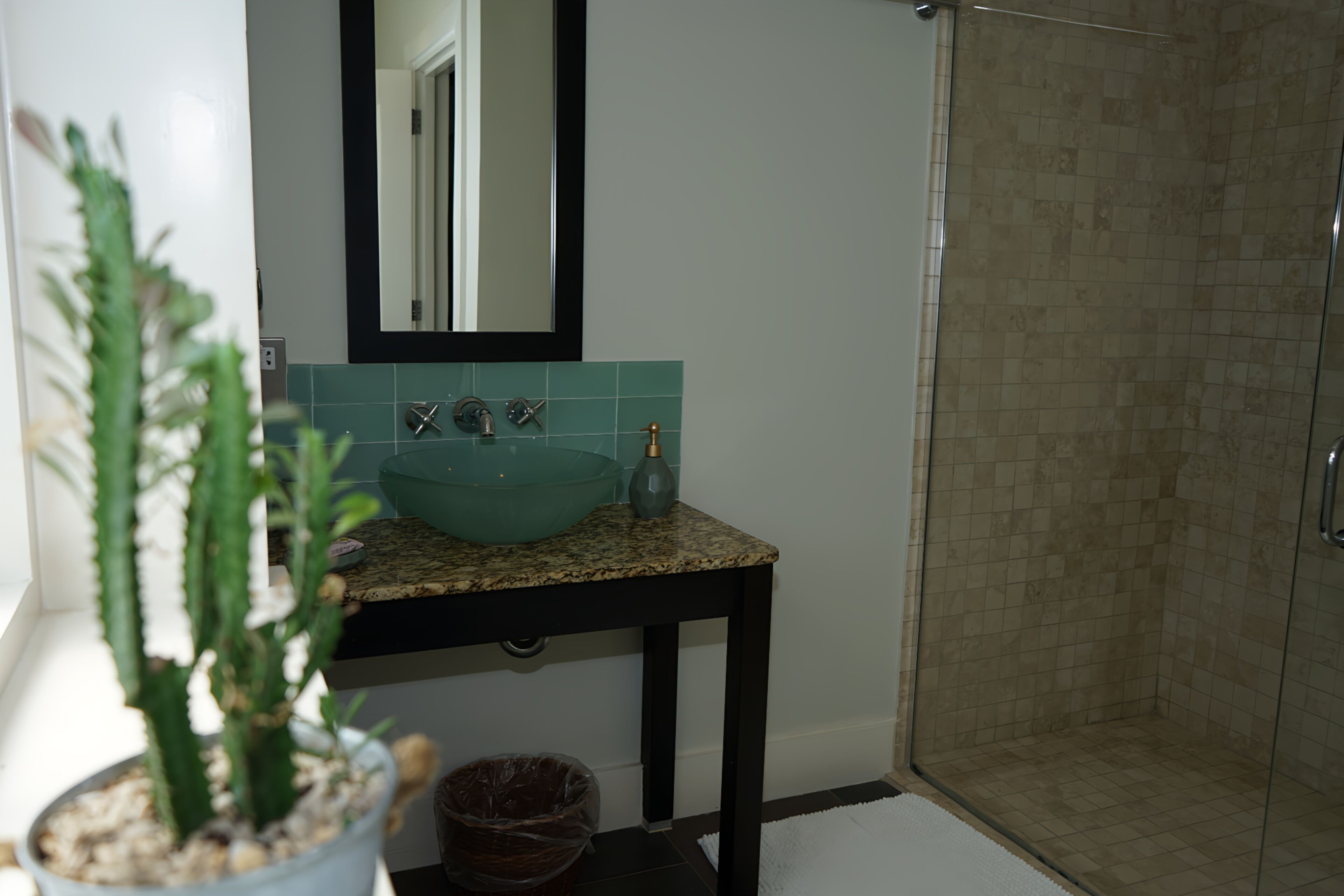 A bathroom with a granite countertop, a teal sink, a mirror, a potted plant, and a shower area.