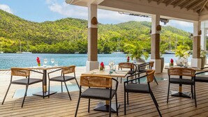 A covered outdoor dining area with tables and chairs, overlooking a body of water and lush green hills.