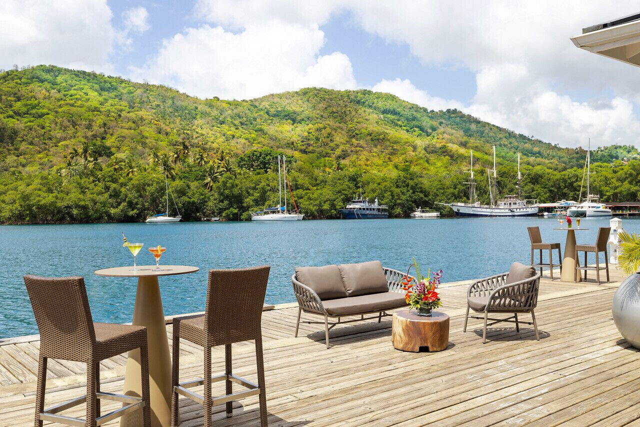 Una terraza de madera con mobiliario de mimbre, una mesa redonda y un sofá con vista a un lago donde se ven barcos y una colina boscosa al fondo.