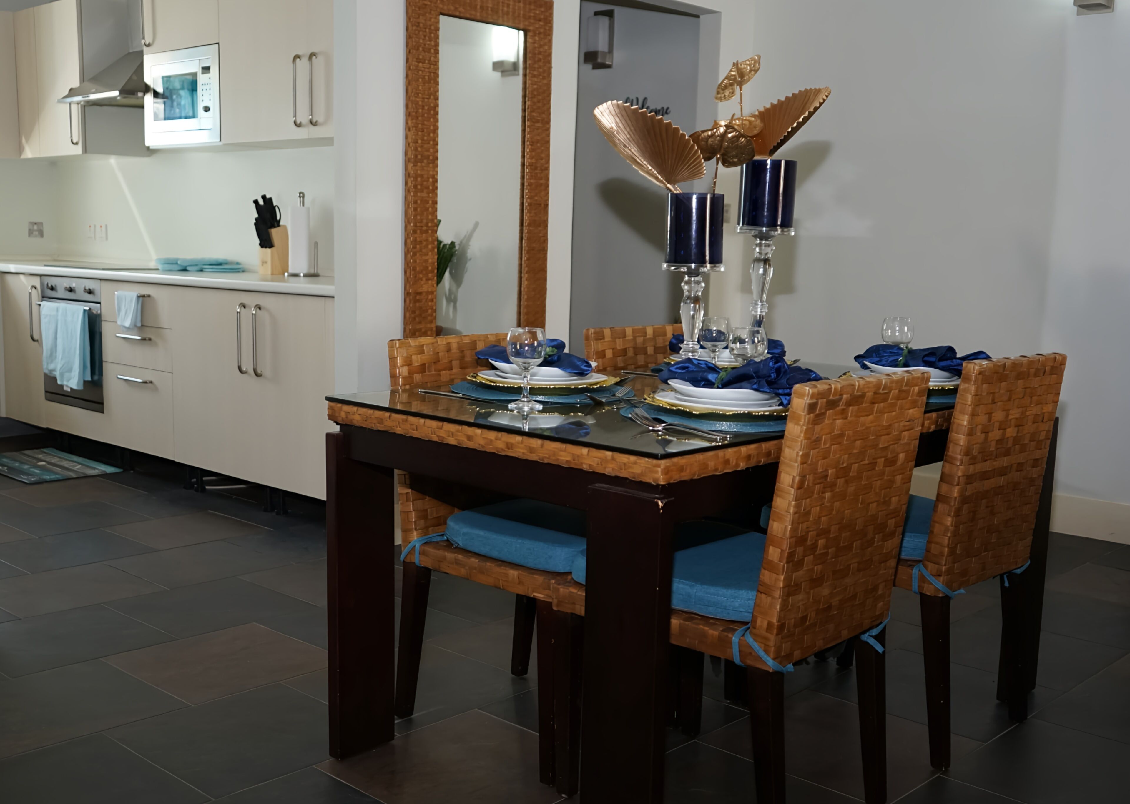 Un comedor con mesa de vidrio dispuesta para cuatro personas, rodeado de sillas de mimbre con cojines azules.
