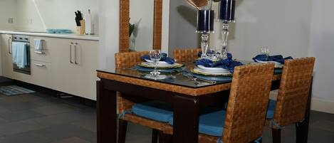 Un comedor con mesa de vidrio dispuesta para cuatro personas, rodeado de sillas de mimbre con cojines azules.