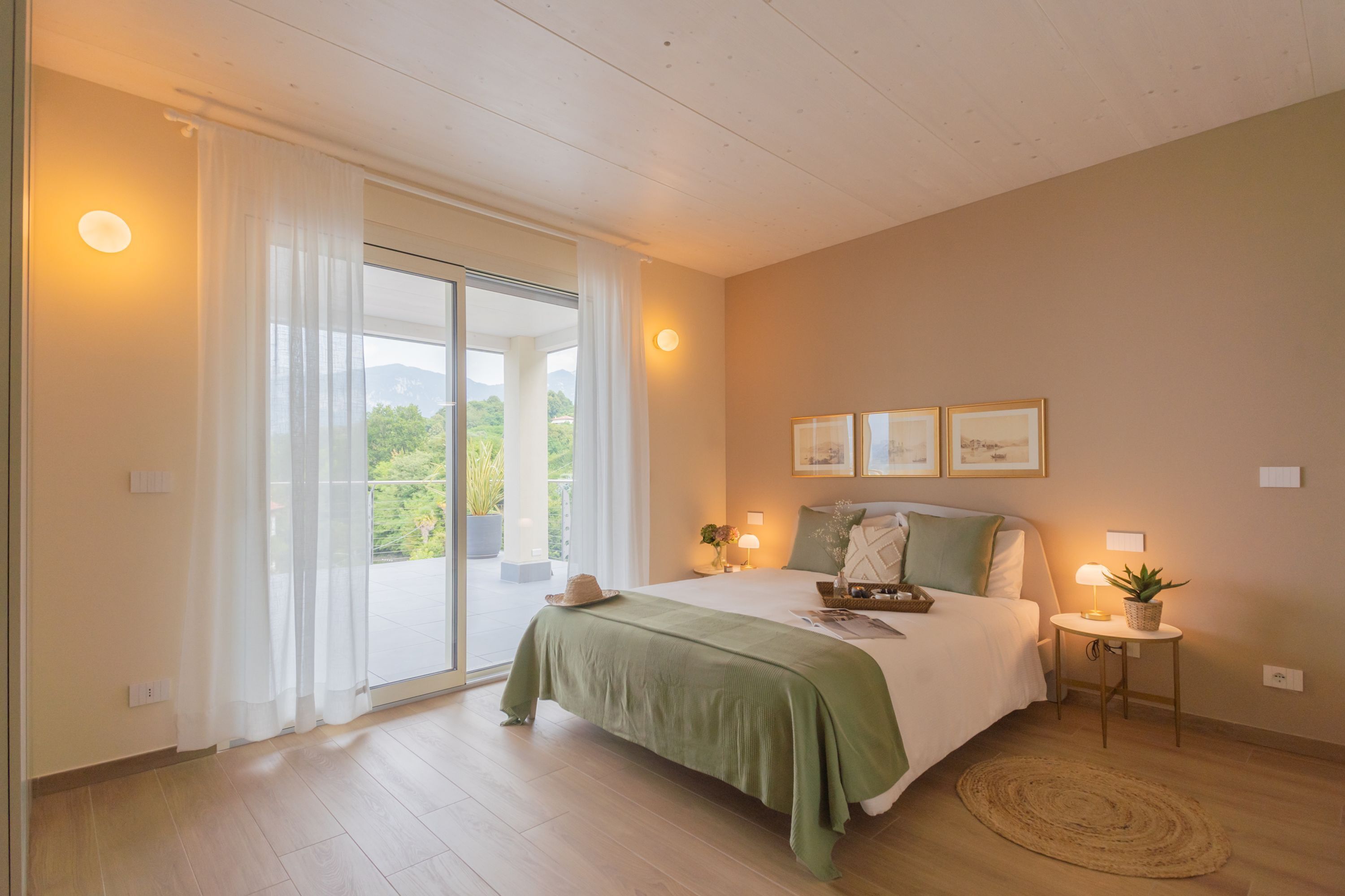 A bedroom with a bed, a bedside table with a lamp, a small potted plant, and a view of greenery through a window.