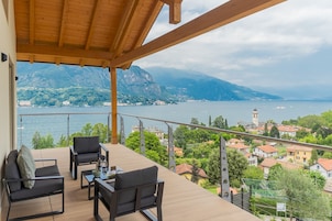 A balcony with a wooden ceiling, furnished with two chairs and a small table, overlooking a lake with mountains in the background.