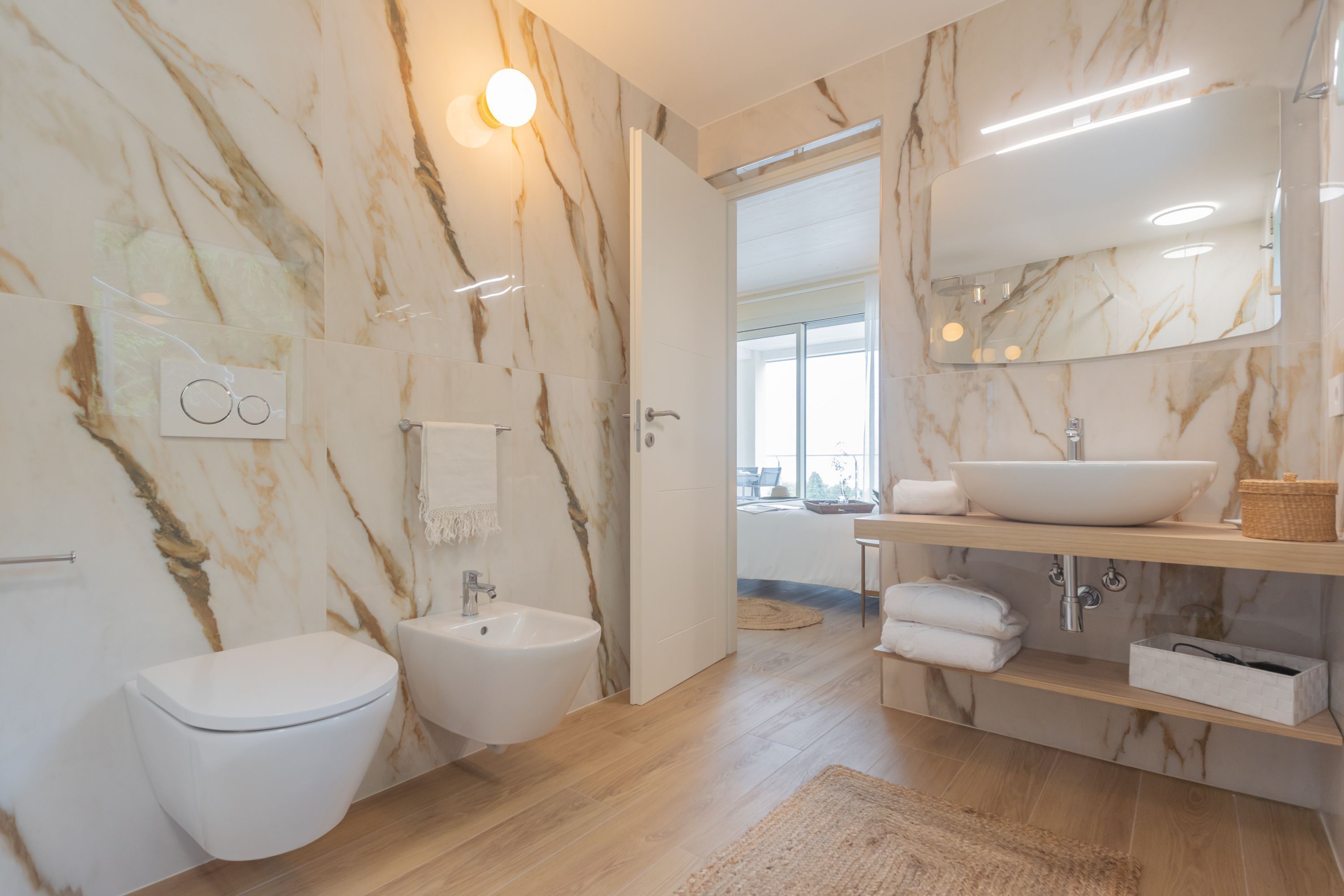A modern bathroom with a marble wall, a bidet, a sink with a large mirror, and a wooden floor.