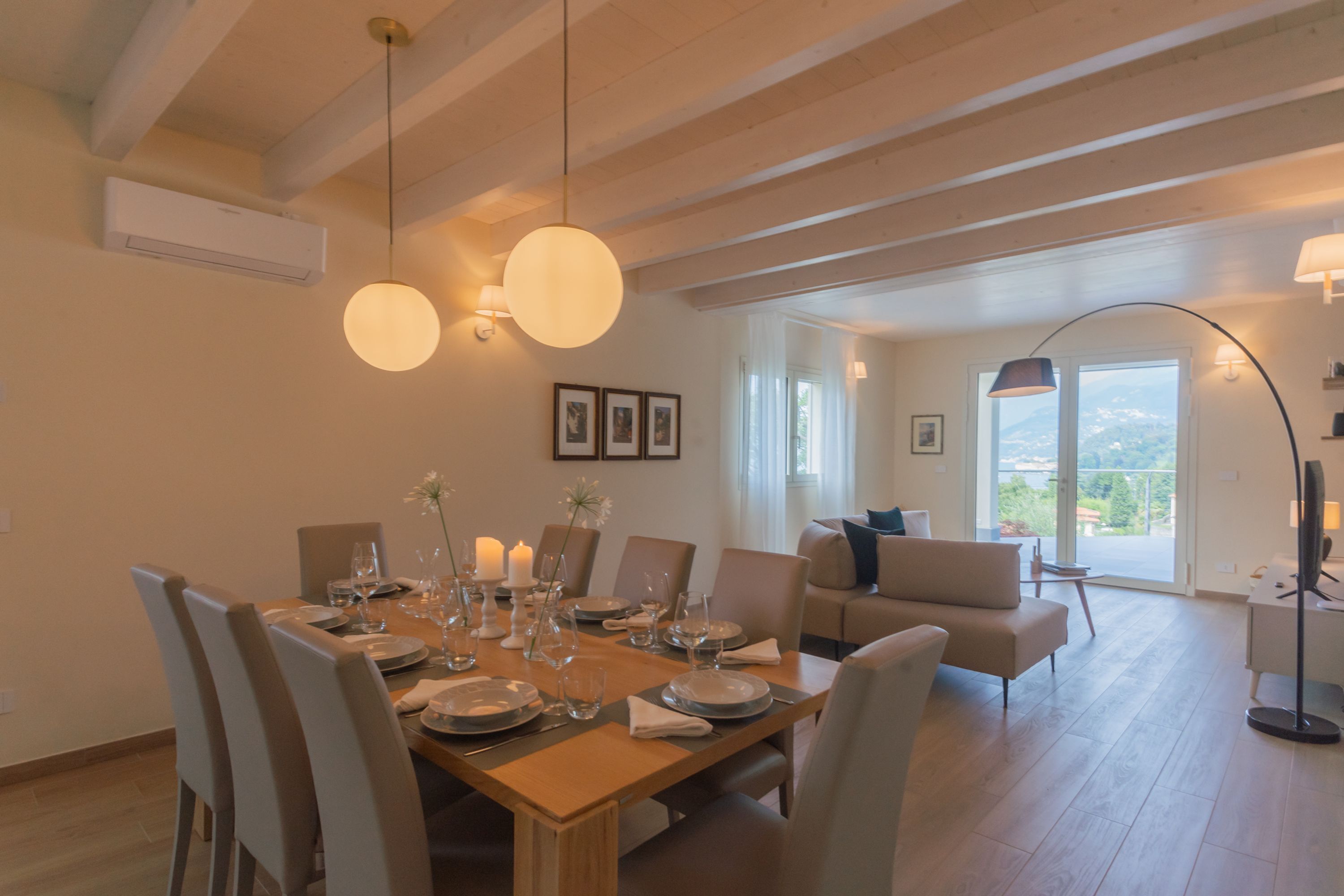 Un comedor con una mesa de madera dispuesta para cuatro personas, rodeada de sillas. Se observa un sofá y una lámpara de pie al fondo.