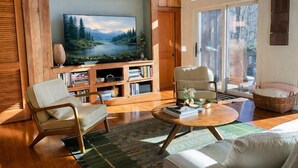A cozy living room with a large flat-screen TV, wooden bookshelves, a comfortable armchair, a round coffee table, and a view of the outdoors through a glass door.