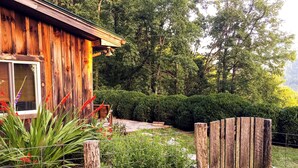 A rustic wooden cabin with a window, surrounded by a garden with various plants and a wooden fence.