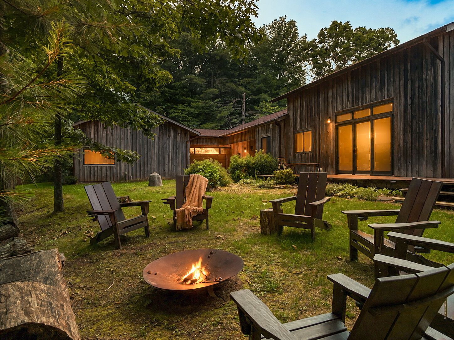 Un espacio al aire libre rústico con sillas Adirondack, un brasero y un edificio de madera con grandes ventanales.