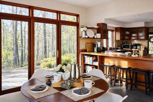 A dining area with a table set for two, a coffee machine, and a view of a forest through large windows. - Mountain Retreat for Two! Japanese Tub. Fast WiFi (Bakersville)