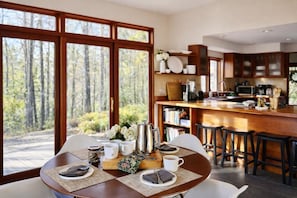 Un coin repas avec une table dressée pour deux personnes, une machine à café et une vue sur une forêt, visible à travers de grandes fenêtres.