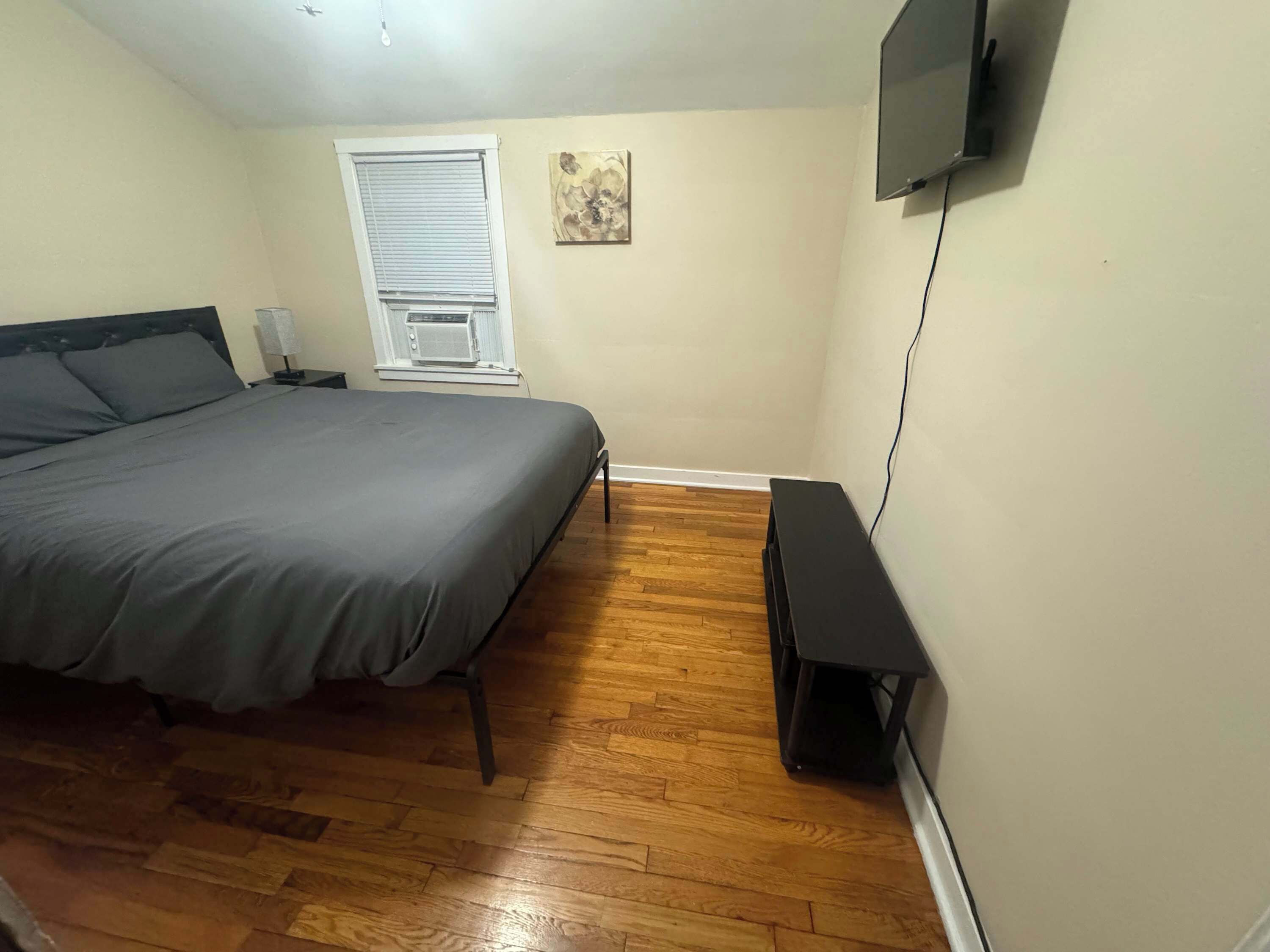 Une chambre à coucher avec un lit, une fenêtre avec des stores, un téléviseur fixé au mur et un plancher en bois.